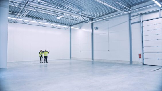 Two people in neon vests and protective headgear stand in an empty warehouse.