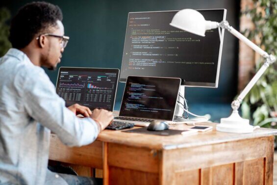 A man sits at his desk and works on his laptop and two monitors.