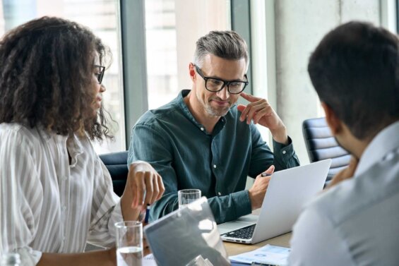 Three diverse business people speak together during a meeting in a boardroom.