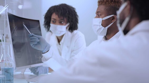 Three doctors in lab coats and masks look at a computer screen.