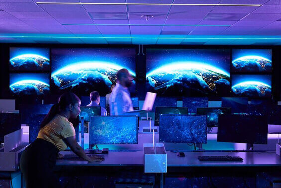 Four people work in a room filled with desktop computers and monitors displaying a view of the Earth from space.