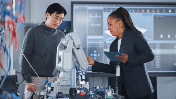 In a science lab, a woman holding a clipboard explains a piece of technical equipment to a young student standing by.