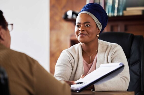 A woman sits at a desk across from someone and smiles as she hands a contract to them to sign.