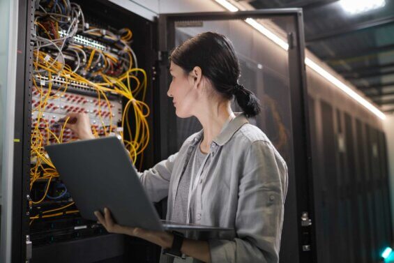 A young woman connects cables in a server cabinet while holding her laptop.