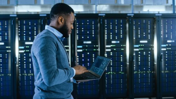 A man consults his laptop as he walks past a row of server cabinets in a data center.