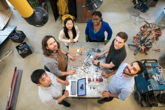 A group of diverse young people stand around a worktable and smile up at the camera.